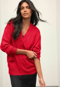 Cardigan rouge avec un col en V, des manches longues et deux poches avant. Texture douce et boutonnière à l'avant. Porté sur une robe noire.