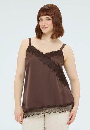 Femme aux cheveux bruns mi-longs portant un caraco en soie marron foncé avec bordure en dentelle et un pantalon de couleur claire, souriant doucement.