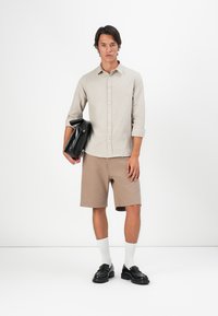 Camisa botão em bege claro, calções ribateados em tom de bege, meias brancas e sapatos pretos. O modelo segura uma bolsa preta, em pé contra um fundo branco.