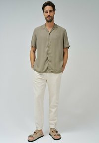 Camisa de botones de manga corta en color verde salvia, combinada con pantalones crema ajustados y sandalias de colores claros. Tejido suave con un ligero brillo.