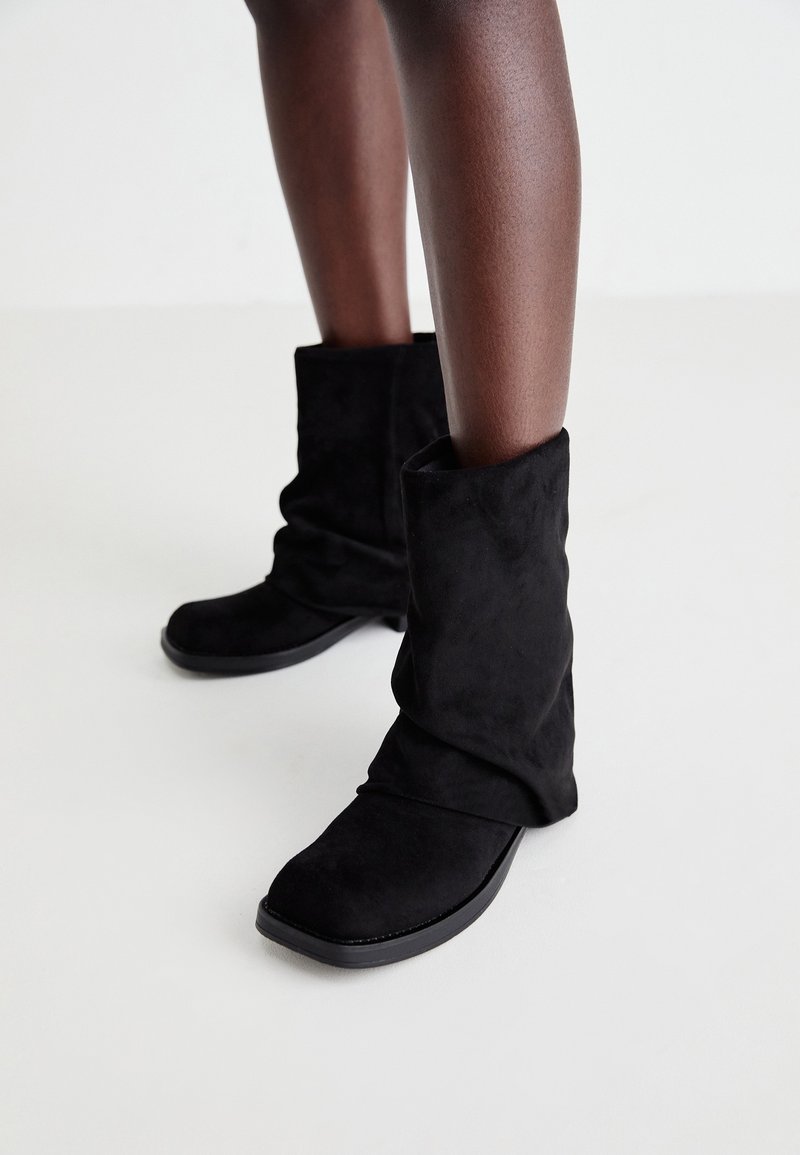 Personne portant des bottes noires en daim souples mi-mollet à bouts carrés, debout sur un sol clair.