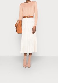 Pull tricot pêche, jupe midi plissée blanche, ceinture marron et bottines roses. Tient un sac en daim marron. Design simple, matériaux texturés.