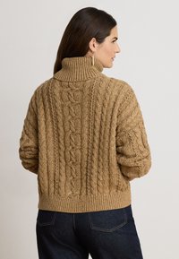 Femme aux longs cheveux foncés portant un pull roulé en maille torsadée beige et un jean bleu foncé, vue de dos sur un fond uni.