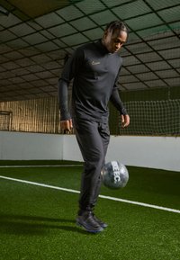 Zwarte sportieve trainingsoutfit met lange mouwen en rits, gedragen door een persoon die een grijze voetbal dribbelt op kunstgras binnen.