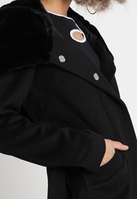Abrigo negro con un suave cuello de pelo sintético negro, grandes botones plateados y bolsillos laterales; el material parece suave con un diseño estructurado.