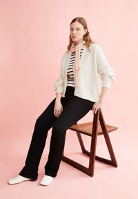White knitted cardigan over a black and white striped shirt, paired with black pants and white shoes, seated on a wooden chair against a pink background.