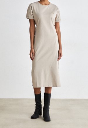 Femme portant une robe midi beige à manches courtes et des bottes noires à talons hauts, debout sur un sol clair contre un mur blanc uni.