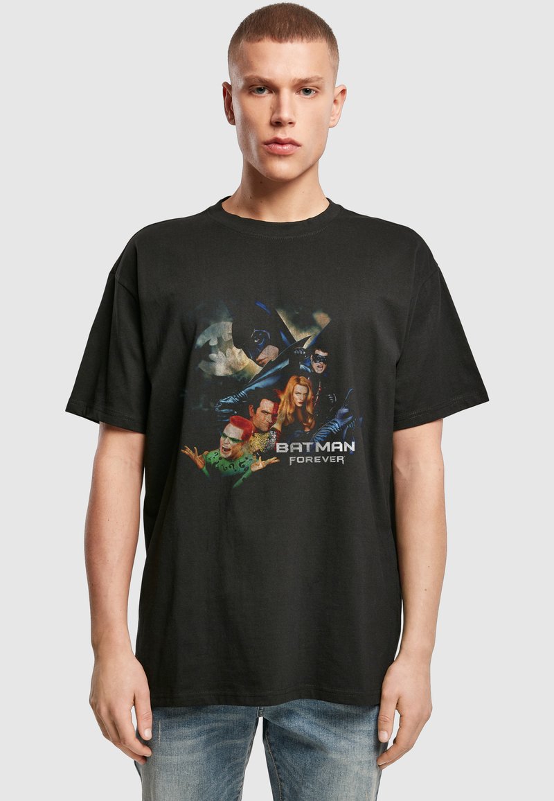Jeune homme portant un T-shirt noir avec un graphisme Batman Forever et un jean bleu, debout devant un fond uni gris clair.