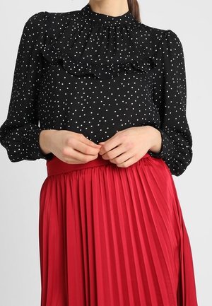 Donna che indossa una camicetta nera a maniche lunghe con pois bianchi e colletto arricciato, abbinata a una gonna plissettata rossa a vita alta.