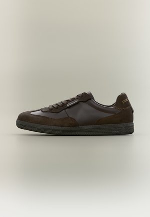 Baskets en cuir et suède marron foncé avec lacets à l'avant, semelle noire texturée et languette de talon marquée sur un fond clair et neutre.