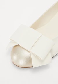 Ballerines avec un embout doré métallisé et un grand nœud blanc, fabriquées à partir de matériaux lisses avec une finition mate.