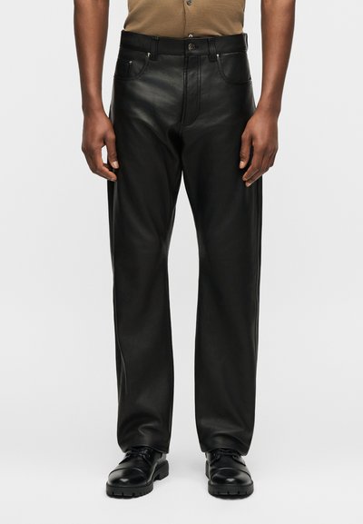 Άνδρας που φοράει μαύρο δερμάτινο παντελόνι με ίσια γραμμή, μαύρα δερμάτινα παπούτσια με κορδόνια και καφέ κοντομάνικο πουκάμισο σε γκρι φόντο.