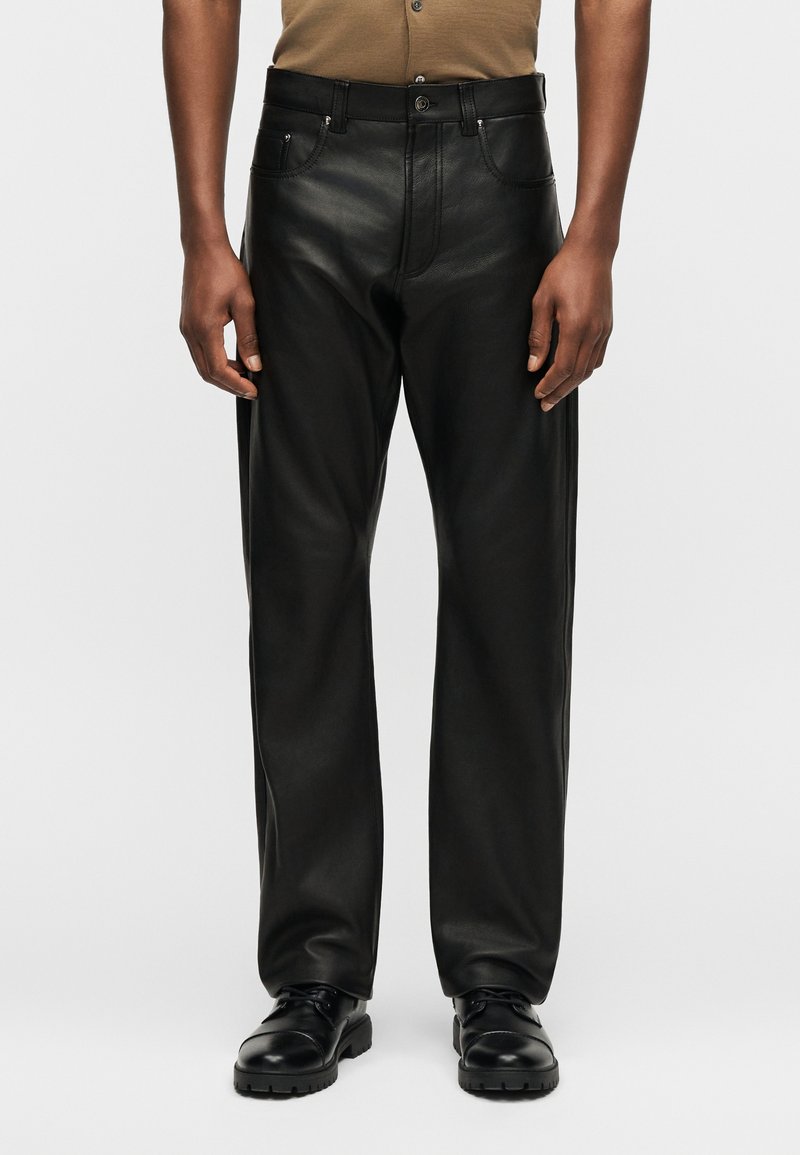 Homme portant un pantalon droit en cuir noir, des chaussures noires à lacets et une chemise marron à manches courtes sur un fond gris.