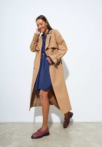 Cappotto beige con un ampio colletto e vita con cintura, indossato sopra un vestito blu navy. Abbinato a mocassini bordeaux su un pavimento chiaro.