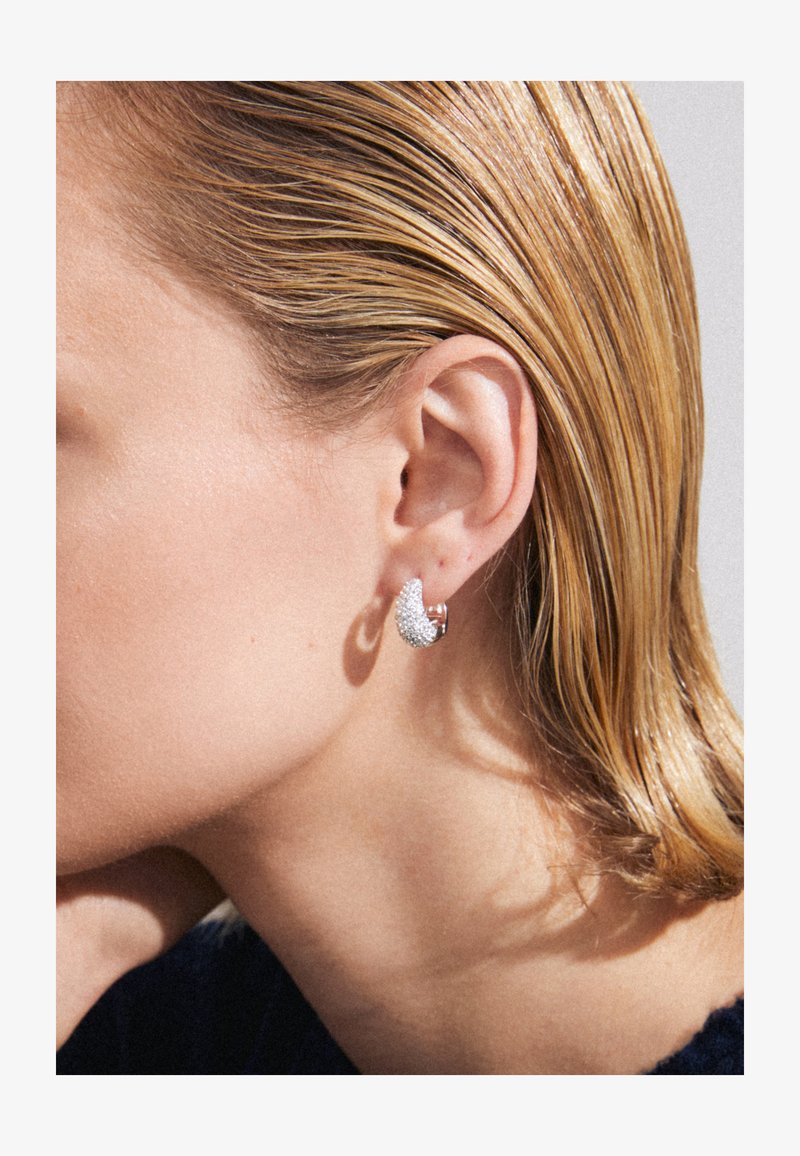 Boucles d'oreilles en ton argent avec un design incurvé et texturé, ornées de petites pierres scintillantes, présentées sur un fond clair.