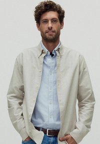 Ljusbeige knäppskjorta lager på lager över en ljusblå och vitrandig skjorta. Matchad med blå jeans och ett brunt bälte. Slät textur.