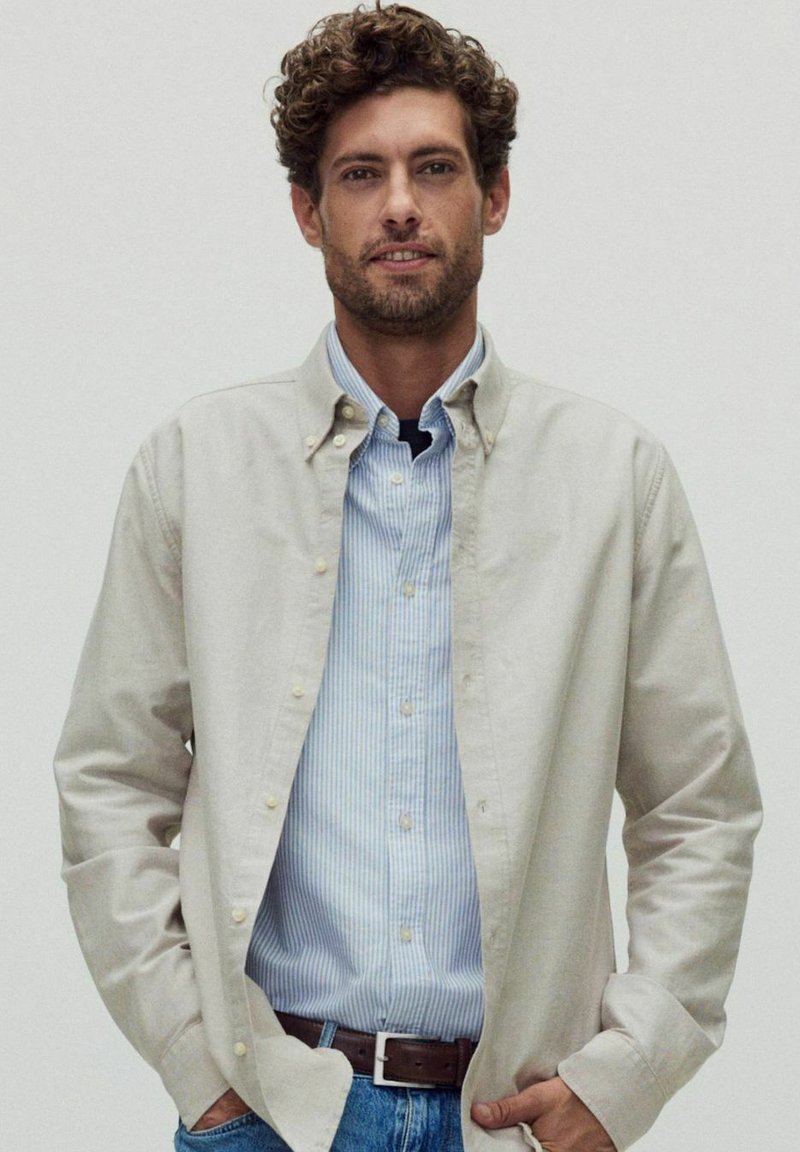 Ljusbeige knäppskjorta lager på lager över en ljusblå och vitrandig skjorta. Matchad med blå jeans och ett brunt bälte. Slät textur.