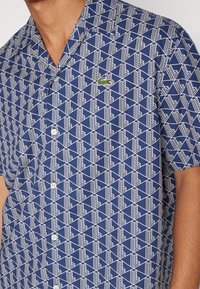Krátký rukáv modré košile s geometrickým bílým vzorem, knoflíky a malým zeleným logem. Vyrobeno z lehkého materiálu s texturovaným povrchem.
