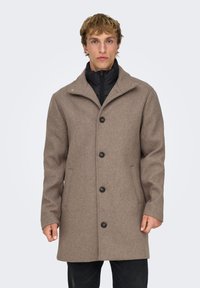 Manteau marron en mélange de laine avec col, cinq boutons, poches latérales et une doublure intérieure noire. Texture lisse et coupe cintrée.