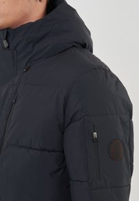 Giubbotto puffer blu scuro con cappuccio, dotato di una tasca sul petto con zip e una patch con logo sulla manica sinistra. Tessuto liscio, design trapuntato.