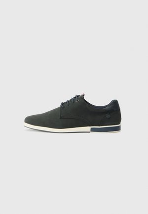 Zapato de gamuza verde oscuro con un acabado suave, punta redondeada, suela bicolor y acentos azules. Presenta perforaciones decorativas y cordones.