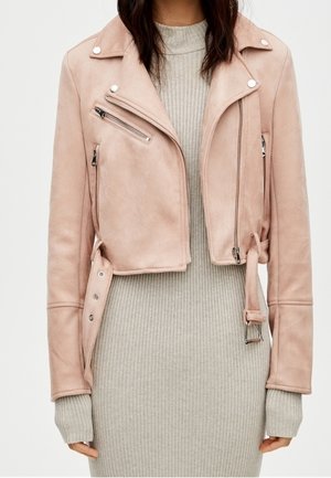 Frau trägt eine hellrosa kurze Jacke mit Reißverschlüssen über einem hellgrauen, gerippten Strickkleid, steht vor einem einfarbigen Hintergrund.