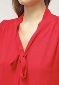 Röd blus med slips vid halsringningen, gjord av lätt tyg. Har mjuka veck vid axlarna och en slät textur.