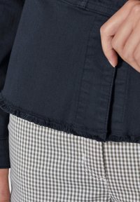 Donker denim top met een rafelige zoom, gecombineerd met geruite broek in olijfgroen en wit. Focus op textuur en contrasterende patronen.