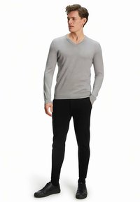 Lichtgrijze v-halssweater gemaakt van gebreide stof, gecombineerd met zwarte slim-fit broek en zwarte sneakers, staande figuur met handen in de zakken.