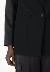 Černý dvouřadý blazer s hladkou texturou, okrouhlými černými knoflíky a ušitými švy, doplněný černými kalhotami s kapsou.