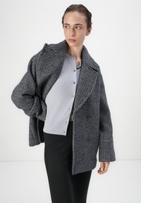 Dobbeltbrystet frakke i grå og sort houndstooth mønster, med en bred krave, tekstureret stof og løs pasform, parret med en grå cardigan.