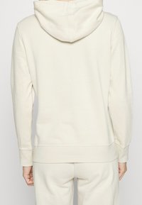 Sweat à capuche beige clair avec une texture douce, manches longues et poignets élastiques. Comprend une poche kangourou à l'avant.