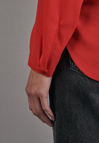 Blouse rouge à manches longues en tissu léger, avec poignets à boutons et ourlet légèrement arrondi, associée à un jean foncé.