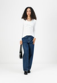 Weißes Langarmshirt mit Strassdesign, kombiniert mit hochtaillierten blauen Denim weit geschnittenen Hosen. Das Model hält eine schwarze Schultertasche.