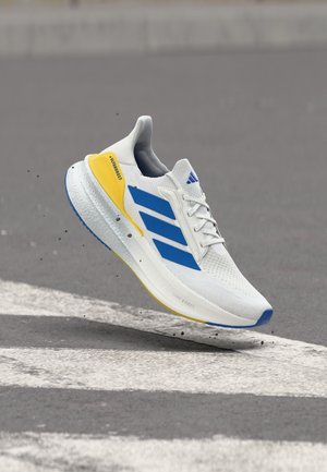 Chaussure de course blanche avec des rayures bleues et un détail jaune au talon, flottant au-dessus d'un bitume gris près de marquages routiers blancs.