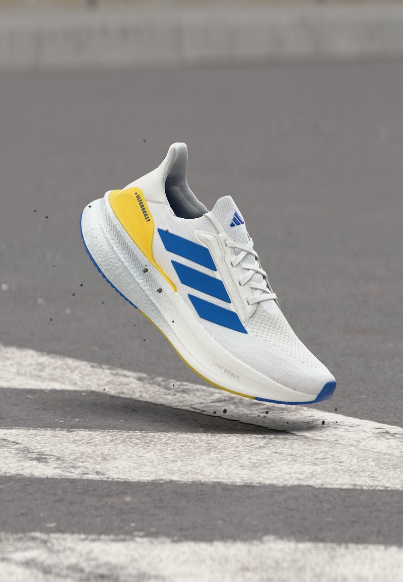 Λευκό αθλητικό παπούτσι με μπλε ρίγες και κίτρινη λεπτομέρεια στη φτέρνα, αιωρούμενο πάνω από γκρι άσφαλτο κοντά σε λευκές σηματοδοτήσεις δρόμου.