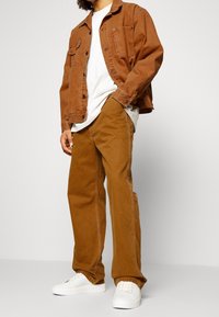 Veste en denim marron avec détails de poches, associée à un pantalon marron et des baskets blanches. La tenue présente une coupe classique et un design à jambes droites.