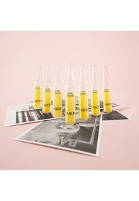 Ocho ampollas transparentes con líquido amarillo etiquetadas como "NUTRI RESTORE" dispuestas sobre fotografías monocromáticas, sobre un fondo rosa.