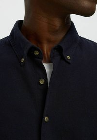 Granatowa koszula zapinana z kołnierzykiem, z dwoma widocznymi mosiężnymi guzikami i teksturowanym materiałem. Koszula jest nałożona na białą podkoszulkę.