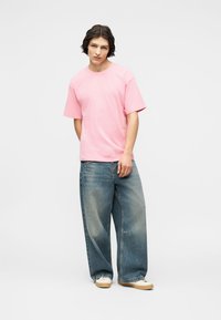 Νεαρό άτομο με κοντά σκούρα μαλλιά, φοράει μια φαρδιά ροζ μπλούζα, φαρδιά μπλε τζιν και μπεζ αθλητικά παπούτσια μπροστά σε ένα απλό λευκό φόντο.