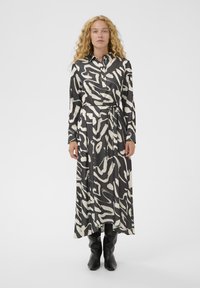 Langes schwarz-weißes Kleid mit abstrakten Mustern, Knopfleiste am Kragen, langen Ärmeln, taillenbündig und fließendem Stoff, kombiniert mit schwarzen Stiefeln.