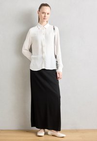 Bluză albă, plisată, cu mâneci lungi și guler, asociată cu o fustă maxi neagră. Modelul poartă pantofi tomate deschise. Fundalul este neutru.