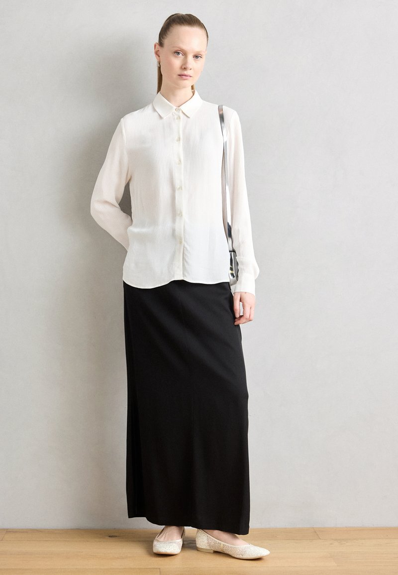 Blouse blanche à manches longues plissées avec un col, associée à une jupe maxi noire. Le mannequin porte des chaussures plates de couleur claire. L'arrière-plan est neutre.