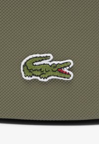 Logo di coccodrillo ricamato di colore verde su uno sfondo testurizzato verde oliva, con contorni bianchi e dettagli rossi sulla bocca.