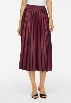 Femme portant une jupe midi plissée bordeaux brillante associée à des escarpins ouverts noirs sur fond blanc.