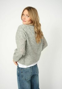 Maglione grigio chiaro, strutturato, con maniche lunghe, scollatura rotonda e vestibilità comoda, indossato sopra una maglietta bianca. Abbinato a jeans blu con logo.
