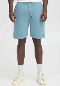 Shorts de trening de culoare albastru deschis, fabricate dintr-un material moale, cu un talie elastică și șnur pentru ajustare. Prezintă buzunare laterale și un mic detaliu sub formă de etichetă neagră.