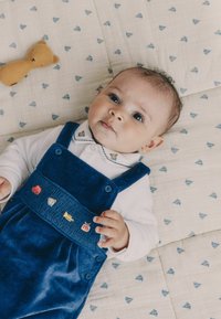 Kék kordbársonyos overallok hímzett állatmintákkal, fehér galléros ing felett viselve. A baba egy mintás textílián fekszik.