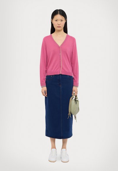Cardigan rosa de botões sobre uma saia midi de ganga escura, combinado com sapatilhas brancas e uma carteira verde, em fundo claro.