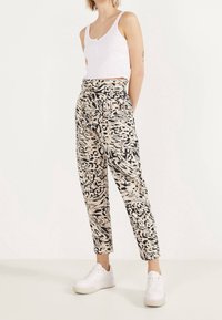 Beige gevoerde broek met een zwart abstract patroon, met een hoge taille en een taps toelopende pijp, gecombineerd met een witte geribbelde tanktop en witte sneakers.